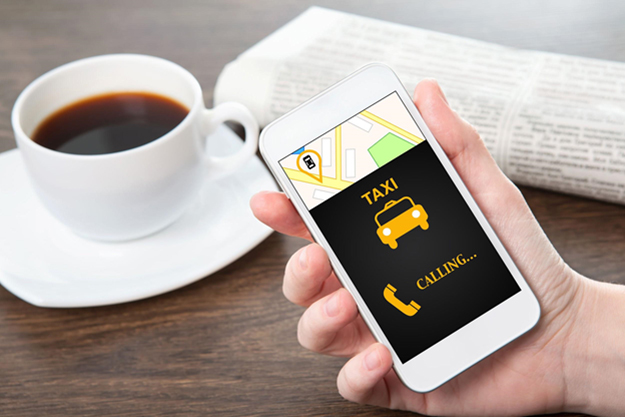 Hand holding a smartphone showing an app used to called a taxi next to a cup of coffee.