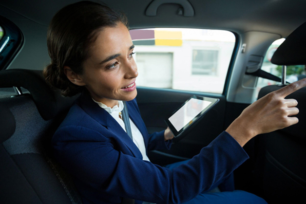 Woman wearing a suit in the backseat of a car and holding a tablet while pointing and given directions towards the driver